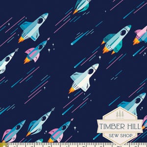 May include: Dark blue fabric with a repeating pattern of cartoon rockets in shades of blue, pink, and white, with orange flames. The rockets are interspersed with shooting stars and small white stars. The fabric has a Timber Hill Sew Shop logo.