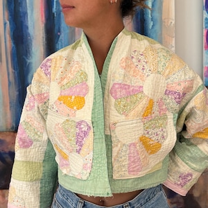 May include: A cropped jacket constructed from quilted fabric featuring a patchwork floral pattern in pastel shades of yellow, pink, and lavender. The garment has a light green trim, a V-neckline, and two front pockets, each adorned with a large flower motif.