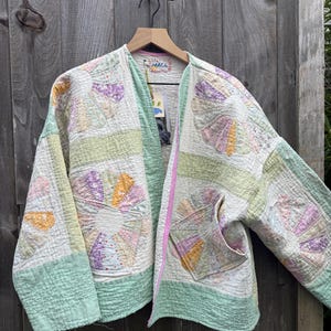 May include: A quilted jacket featuring a patchwork design of fan-shaped patterns in pastel shades of yellow, pink, and lavender on a white background. The garment includes light green border panels, a pink trim along the front opening, and a label reading MACH.