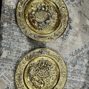 May include: Two antique brass plates with intricate embossed designs. Each plate features a central motif of a fruit basket surrounded by ornate floral and scrollwork borders. The plates have a warm, golden hue.