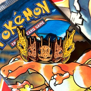 May include: A gold-colored enamel pin featuring a group of three Charizard Pokemon heads from the Pokemon Trading Card Game. The pin is set against a background of a Pokemon Trading Card Game booster pack featuring a Charizard.