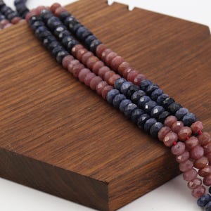 May include: Close-up of three strands of faceted gemstone beads. The beads alternate between deep blue and reddish-brown hues. The strands are displayed on a wooden surface, showcasing the color and texture of the beads. The beads are strung on a red thread.