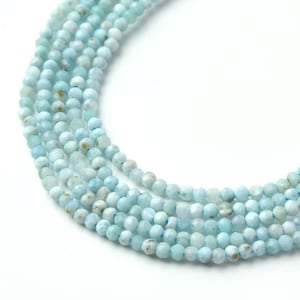 May include: Strands of light blue gemstone beads. The beads are small, round, and faceted, with a pale blue color and subtle variations. The beads are strung together, creating a flowing, organic shape. The background is white.