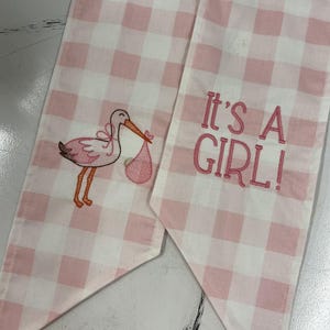 Peut inclure: Tissu vichy rose et blanc avec une cigogne brodée portant un bébé dans un ballot rose. Le texte "It's a GIRL!" est brodé en rose. Un article décoratif pour une baby shower.