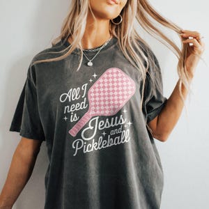 May include: A dark grey t-shirt with a pink pickleball paddle graphic and the text "All I need is Jesus and Pickleball".