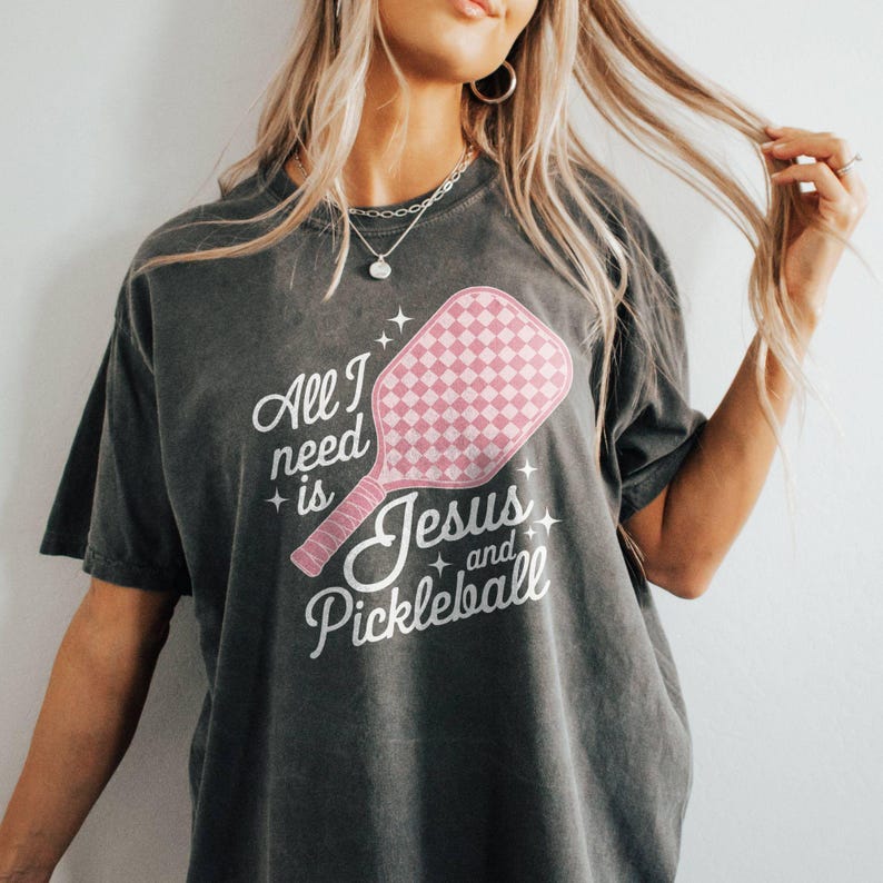 May include: A dark grey t-shirt with a pink pickleball paddle graphic and the text "All I need is Jesus and Pickleball".