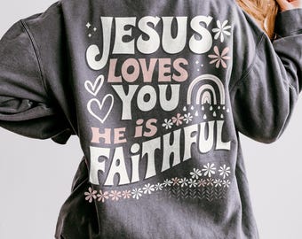 May include: A dark gray sweatshirt with the text "Jesus Loves You He is Faithful" in white and pink lettering. The text is in a retro font style and is surrounded by small flowers and a rainbow.