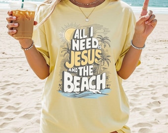 May include: A pale yellow short-sleeve t-shirt featuring a graphic print with a sun, palm trees, and ocean waves. The text reads, All I NEED is JESUS AND THE BEACH. The design is centred on the chest in dark brown and cream lettering.
