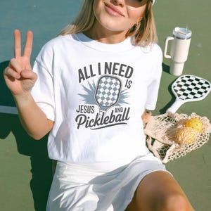 May include: White t-shirt with the text "All I need is Jesus and Pickleball" with a graphic of a pickleball paddle.
