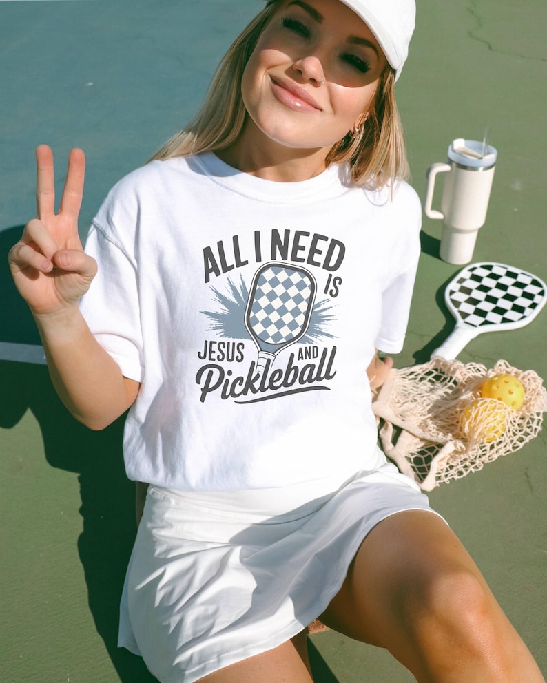 May include: White t-shirt with the text "All I need is Jesus and Pickleball" with a graphic of a pickleball paddle.