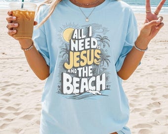 May include: A light blue t-shirt featuring a graphic print with a yellow sun, palm trees, and the text "ALL I NEED IS JESUS AND THE BEACH" in white and yellow lettering. The design includes stylized line work depicting a coastal scene.