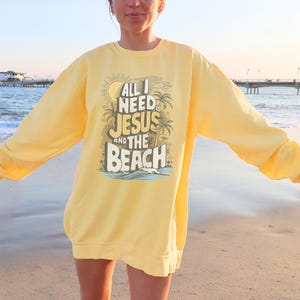 Op de afbeelding: Geel sweatshirt met de tekst "All I Need is Jesus and the Beach" in een grafisch ontwerp. Het sweatshirt heeft lange mouwen en een ronde hals. De achtergrond is een strand met een pier en de zee.