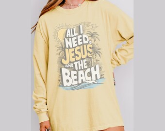 May include: A pale yellow long-sleeved shirt featuring the text "ALL I NEED IS JESUS AND THE BEACH" in a graphic design with a sun, palm trees, and waves. The shirt is a casual, comfortable style.