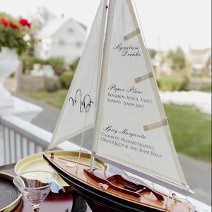 May include: A model sailing boat with a wooden hull and white fabric sails. The sails feature black printed text reading Signature Drinks, Paper Plane, and Spicy Margarita, along with a monogram. The boat is mounted on a black stand.