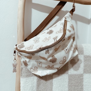 May include: A white fanny pack with a brown strap and a zipper closure. The fanny pack is decorated with a pattern of brown Mickey Mouse heads, pretzels, popcorn buckets, ice cream cones, and churros.