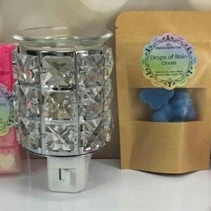 Puede incluir: Un calentador de cera eléctrico de plata y cristal con una base enchufable. Se muestran tres fundidos de cera en la imagen: uno naranja, uno rosa y uno blanco. También se muestra una bolsa de papel marrón con fundidos de cera azules y el texto "Drops of Rain Clouds". Un frasco transparente con fundidos de cera rosas está en el fondo.