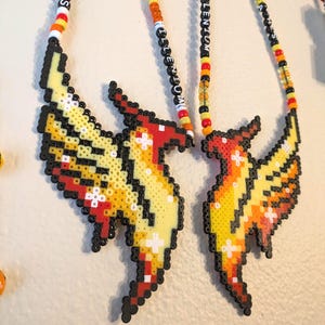 May include: Two beaded necklaces featuring pixelated phoenix designs in shades of yellow, orange, red, and black. The necklaces have beaded strands with the text "FROM THE ASHES" and "MILLENNIUM".