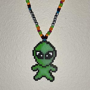 May include: A green alien-shaped beaded charm hangs from a colorful beaded necklace with the words "BABE" and "SPACE" on either side.