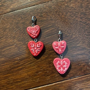 Puede incluir: Dos encantos de corazón rojo con diseños florales blancos. Cada encanto está unido a un clip plateado.
