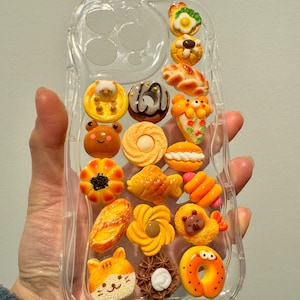 May include: A clear phone case with various colourful 3D food charms attached to it. The charms include doughnuts, bread, fish, and other baked goods.