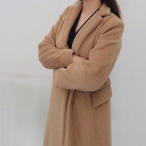 May include: A long, brown, fuzzy coat with a collar and a button closure. The coat is worn by a person with their arms crossed.