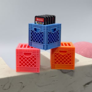 May include: Three miniature plastic crates, one blue, one orange, and one pink, stacked on a white surface. The blue crate has a stack of Nintendo Switch game cartridges inside.