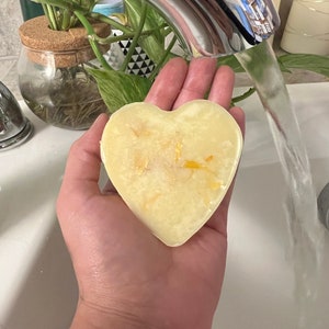 May include: A heart-shaped bar of yellow soap with visible flecks, held under running water. The soap is in a person's hand, with a tattoo visible on the wrist. The background includes a faucet, a sink, and a plant in a glass jar.