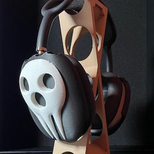 Pu&ograve; includere: Cuffie nere con un designo a forma di teschio bianco sugli auricolari, appoggiate su un supporto in legno.