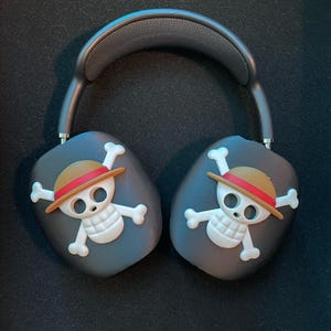May include: Black wireless headphones with a skull and crossbones design featuring a straw hat and red band. The skull is white with a black outline.