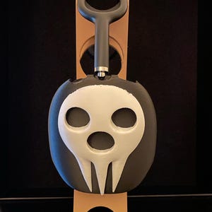 Pu&ograve; includere: Un supporto per cuffie a forma di teschio nero e bianco con un design di teschio bianco. Il supporto &egrave; in plastica e ha una base nera.