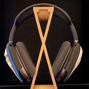 Pu&ograve; includere: Un supporto per cuffie in legno con finitura marrone chiaro. Il supporto ha la forma di una X e ha un piccolo bordo superiore per tenere le cuffie. Un paio di cuffie nere con dettagli bianchi sono appoggiate sul supporto.