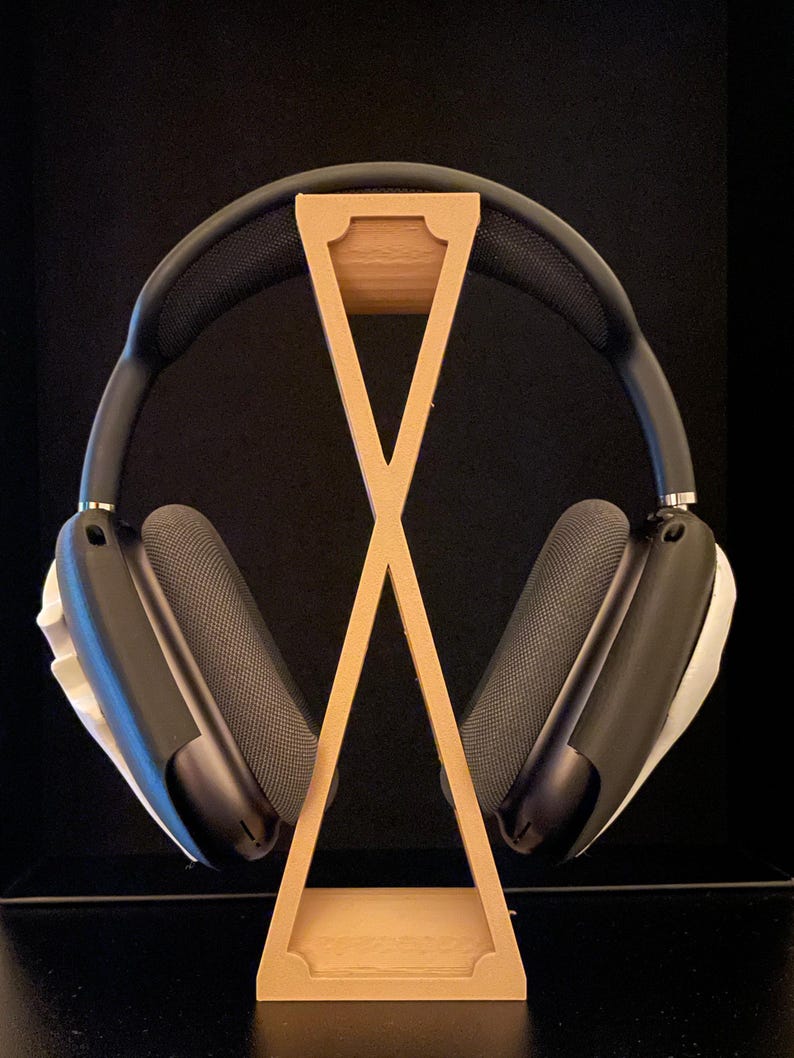 Pu&ograve; includere: Un supporto per cuffie in legno con finitura marrone chiaro. Il supporto ha la forma di una X e ha un piccolo bordo superiore per tenere le cuffie. Un paio di cuffie nere con dettagli bianchi sono appoggiate sul supporto.