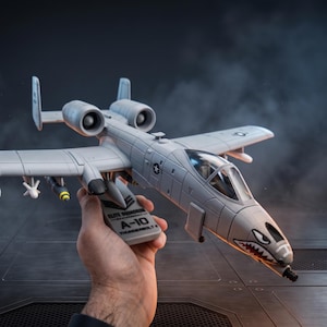 May include: A gray A-10 Thunderbolt II model airplane with detailed features, including missiles and a shark mouth design on the nose. The model is held in a hand against a dark, industrial-style background.