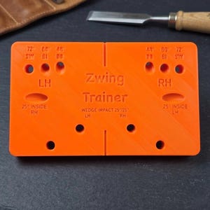 May include: Bright orange "Zwing Trainer" tool with text and markings for wedge impact and angles. Features holes and slots for various uses. A woodworking chisel and leather are in the background.