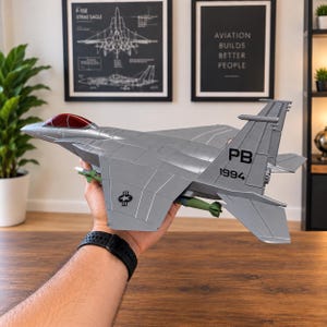 May include: A silver model fighter jet with a red cockpit canopy, green missiles mounted under the wings, and the black markings PB 1994 on the vertical stabilizer. The aircraft features a star and bar insignia on the wing and a sleek design.