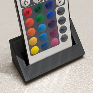 May include: A black remote control with a white face and a variety of buttons. The buttons are labeled with letters, numbers, and words like "ON", "OFF", "FLASH", "STROBE", "FADE", and "SMOOTH". The remote is sitting in a black stand.