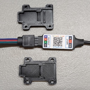 May include: An overhead shot shows two black 3D-printed mounting brackets and an LED controller. The controller has a white label with the text "OUTPUT BRG V+" and "INPUT VCC V-". The controller has wires with red, green, blue, and black insulation.