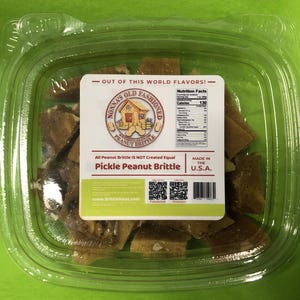 May include: A clear plastic container filled with pieces of peanut brittle. The label reads "Nonna's Old Fashioned Peanut Brittle" and "Pickle Peanut Brittle." The label also includes nutritional information and states "Made in the U.S.A."