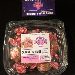May include: A clear plastic container filled with caramel cremes wrapped in red and white wrappers. The container has a label that reads "Caramel Cremes" and "www.Space-Candy.Shop". The label also includes a QR code and nutritional information. The container is sitting on a black surface.