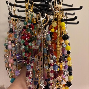 May include: A collection of beaded lanyards and keychains hanging from a black rotating display stand. The accessories feature a variety of colourful glass, plastic, and metal beads in yellow, red, blue, and black, attached to gold and silver metal clasps.