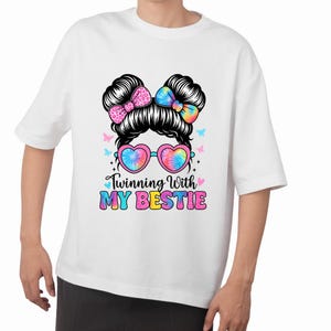 May include: White t-shirt featuring a graphic of a woman's hairstyle with heart-shaped sunglasses and bows. The design includes the text "Twinning With My Bestie" in colorful lettering. Butterflies and tie-dye patterns add to the design.