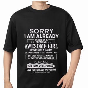Può includere: T-shirt nero con testo bianco. Il testo recita "SORRY I AM ALREADY TAKEN BY A FREAKING AWESOME GIRL" e include frasi sul partner di chi lo indossa. La maglietta è a girocollo.