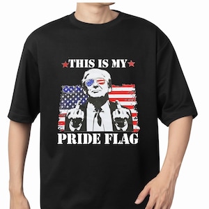 May include: Black t-shirt featuring a graphic of a man with sunglasses in the colors of the American flag, giving the middle finger. The text reads "THIS IS MY PRIDE FLAG" in white.