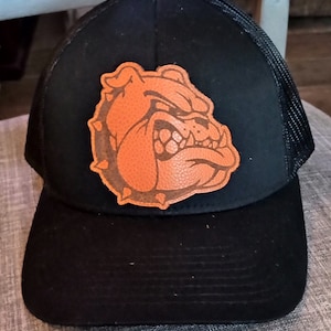 Può includere: Cappellino da camionista nero con una toppa di bulldog in pelle marrone. Il bulldog ha un'espressione ringhiante con un design in pelle testurizzata.