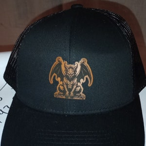 Könnte beinhalten: Schwarze Trucker-Kappe mit einem braunen gestickten Gargoyle-Design.