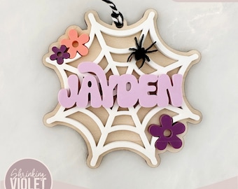 Può includere: Un ornamento in legno a forma di ragnatela con il nome "Jayden" in lettere rosa, un ragno nero e fiori viola e arancioni. L'ornamento è tagliato al laser ed è pronto per essere assemblato.