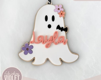May include: A white ghost with a pink and purple flower crown and the name "Layla" written in pink. The ghost has a black bat on its side. This is a laser-cut SVG file for crafting.