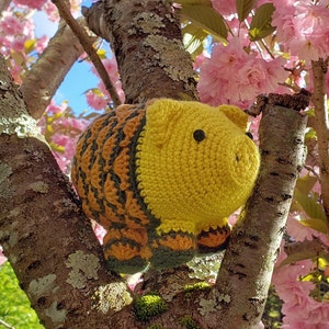 Op de afbeelding: Een geel gehaakt varken met bruine en groene strepen zit in een tak van een boom met roze bloesems.