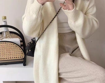 May include: A white long-sleeved cardigan with a V-neckline. The cardigan is made of a soft, knitted fabric and has a relaxed fit. The model is wearing the cardigan with a white tank top and beige pants.