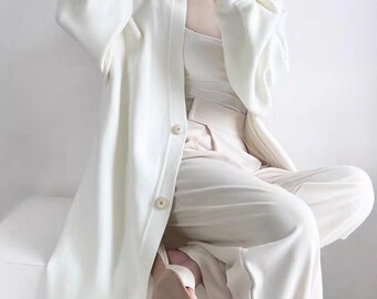May include: A white long-sleeved cardigan with a button closure and a relaxed fit. The cardigan is worn over a white tank top and cream-colored pants.